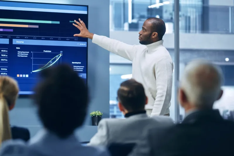Man leading corporate meeting discussing charts on wall monitor.